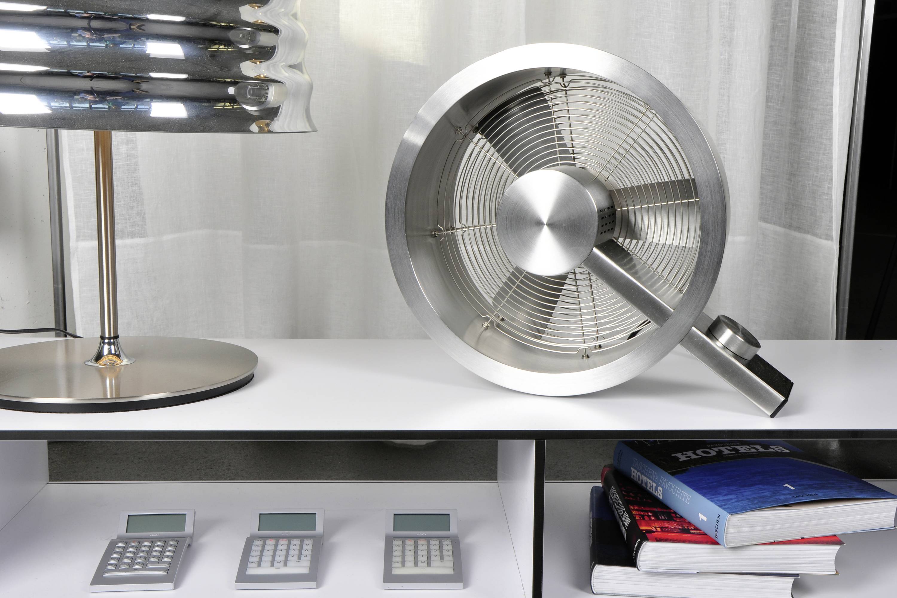 Ein silberner Ventilator und eine Lampe stehen auf einem Tisch. Darunter liegen Bücher und drei Taschenrechner auf einem Regal.
