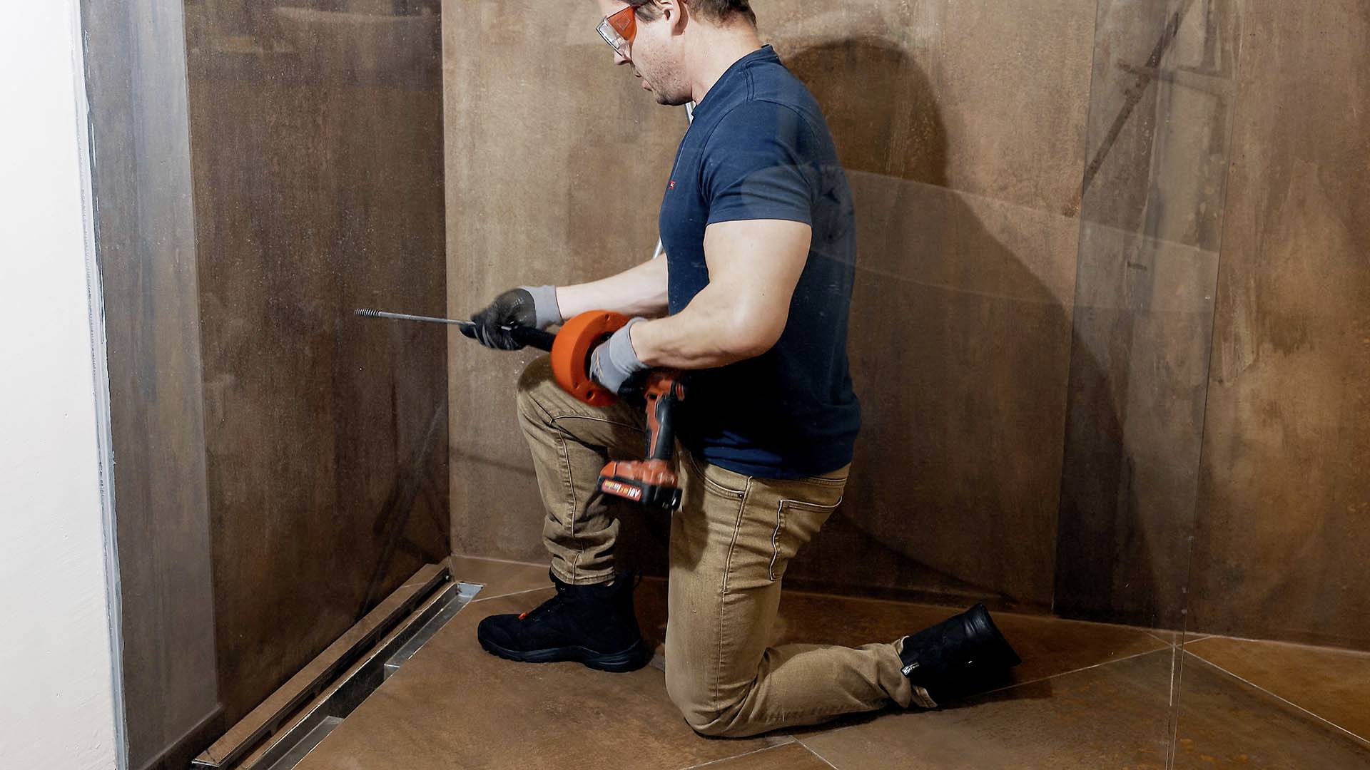 Ein Handwerker kniet auf einem Boden und verwendet eine elektrische Bohrmaschine, um eine Schraube in eine Wand zu befestigen.