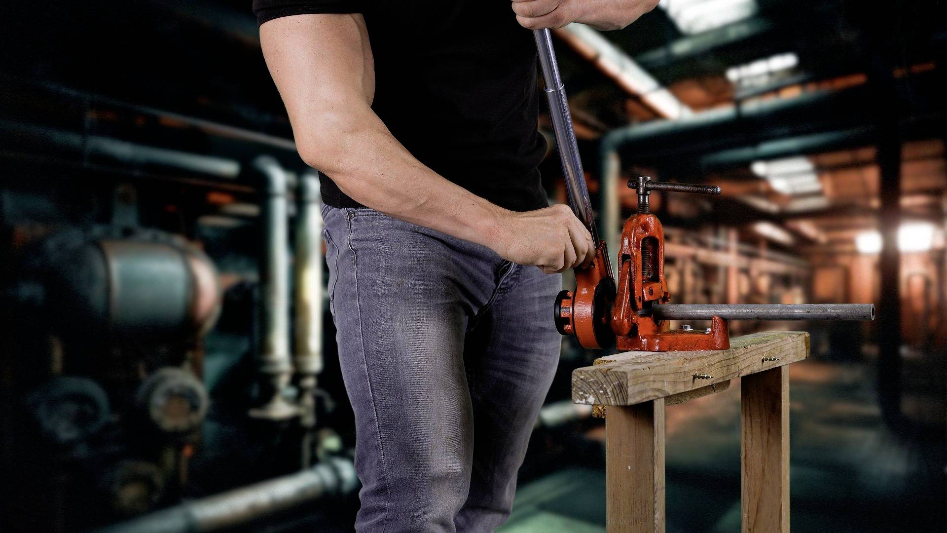 Eine Person bedient ein Rohrbiegewerkzeug in einer Fabrikumgebung. Im Hintergrund sind Rohre und industrielle Maschinen sichtbar.