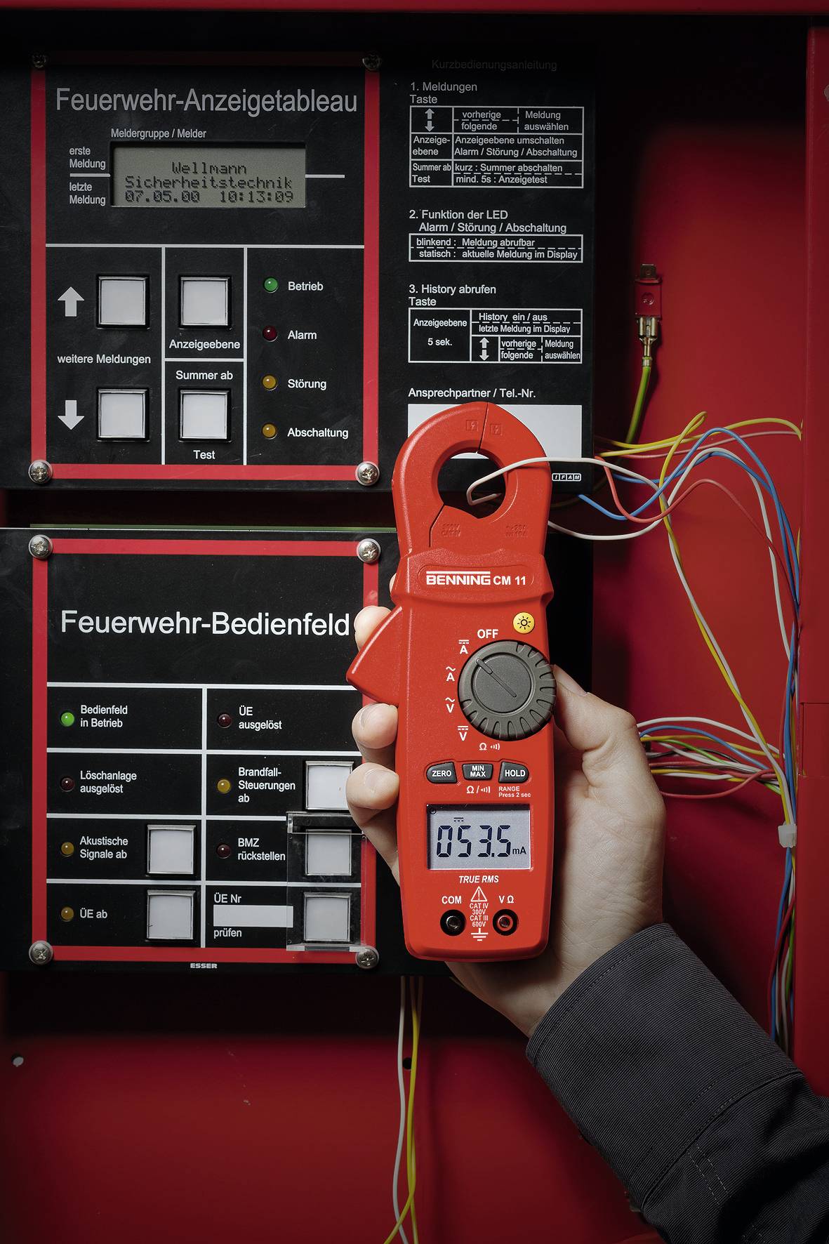 Eine Person hält ein Spannungsmessgerät vor einer Schalttafel der Feuerwehr, auf der Anzeigen und Bedienelemente sichtbar sind.