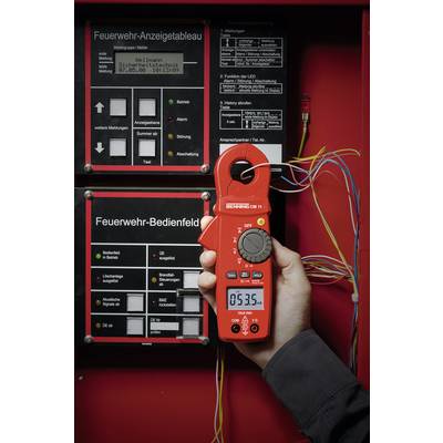 Eine Person hält ein Spannungsmessgerät vor einer Schalttafel der Feuerwehr, auf der Anzeigen und Bedienelemente sichtbar sind.
