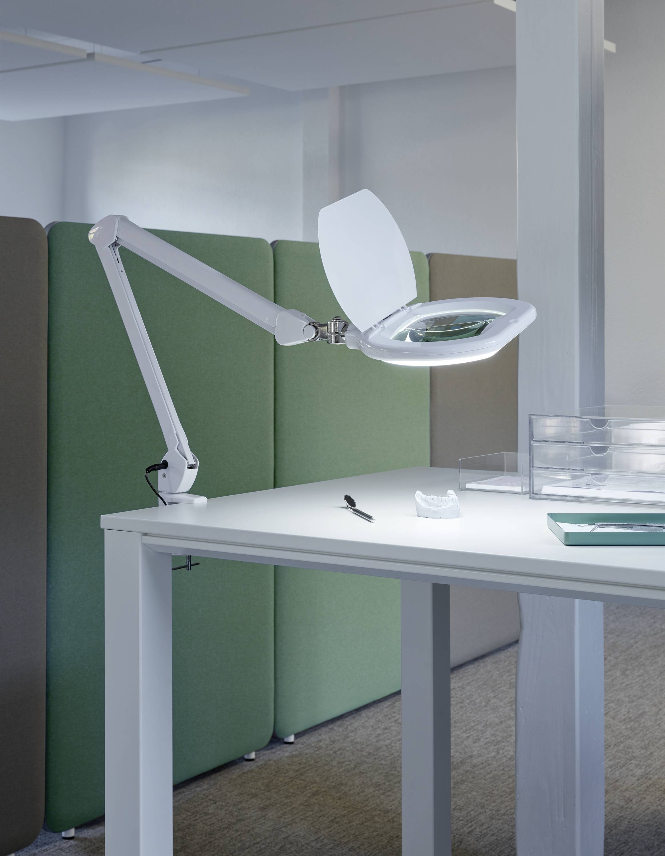 Ein heller Arbeitsplatz mit einer verstellbaren Lupenlampe über einem Tisch. Im Hintergrund stehen grüne Trennwände.