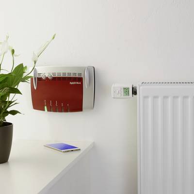 Ein Wifi-Router hängt an der Wand neben einem Thermostat auf einem Heizkörper. Eine Pflanze steht auf dem Tisch daneben.