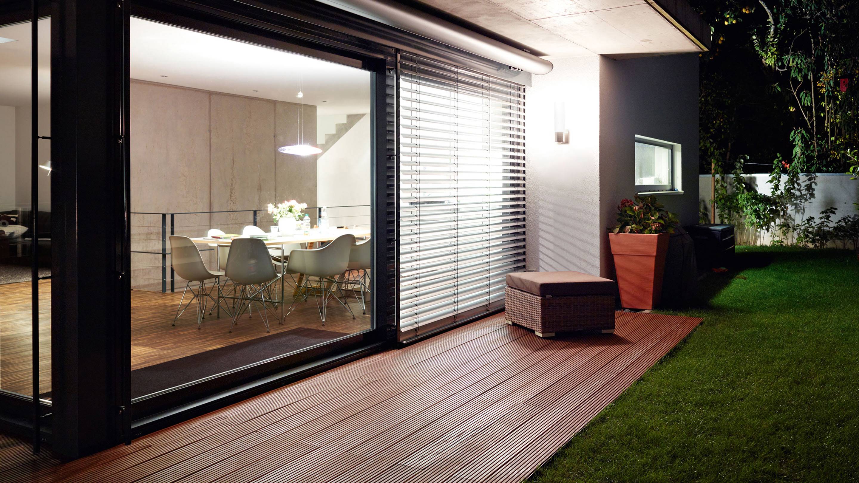 Ein modernes Wohnzimmer mit Glasfront, das in einen beleuchteten Garten mit Holzterrasse und Rasen übergeht.