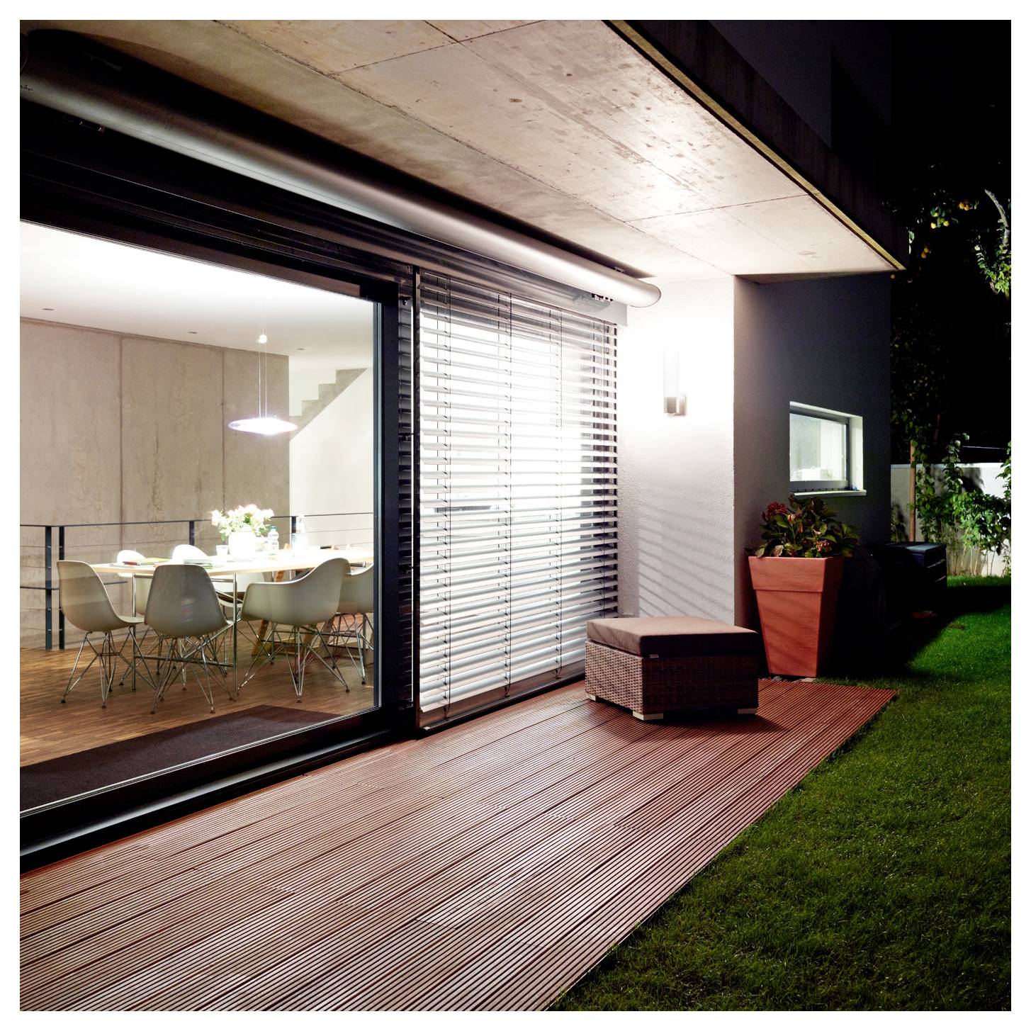 Modernes Haus-Terrassenzimmer bei Nacht mit Glasschiebetüren, Holzterrasse, Außensitzbereich und beleuchteter Essfläche im Inneren.