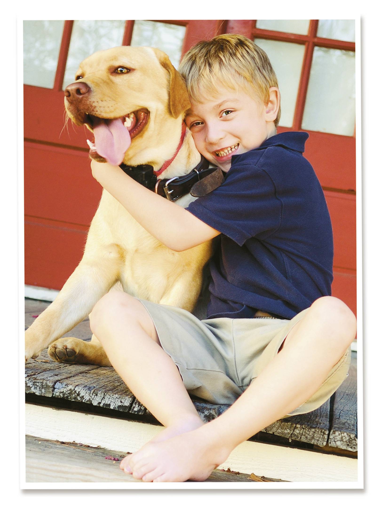 Ein Junge sitzt lächelnd neben einem großen, gelben Hund und umarmt ihn herzlich vor einer roten Holztür.