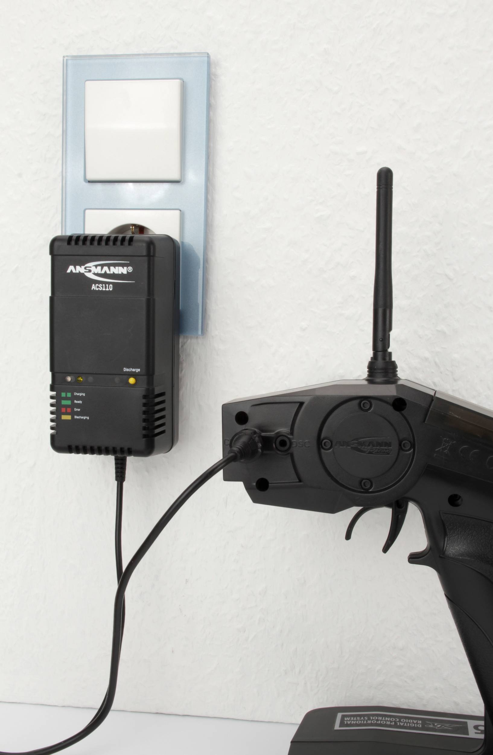 Fernsteuerung lädt an einer Wandsteckdose mit Ansmann-Ladegerät. LED-Anzeigen zeigen den Ladevorgang.