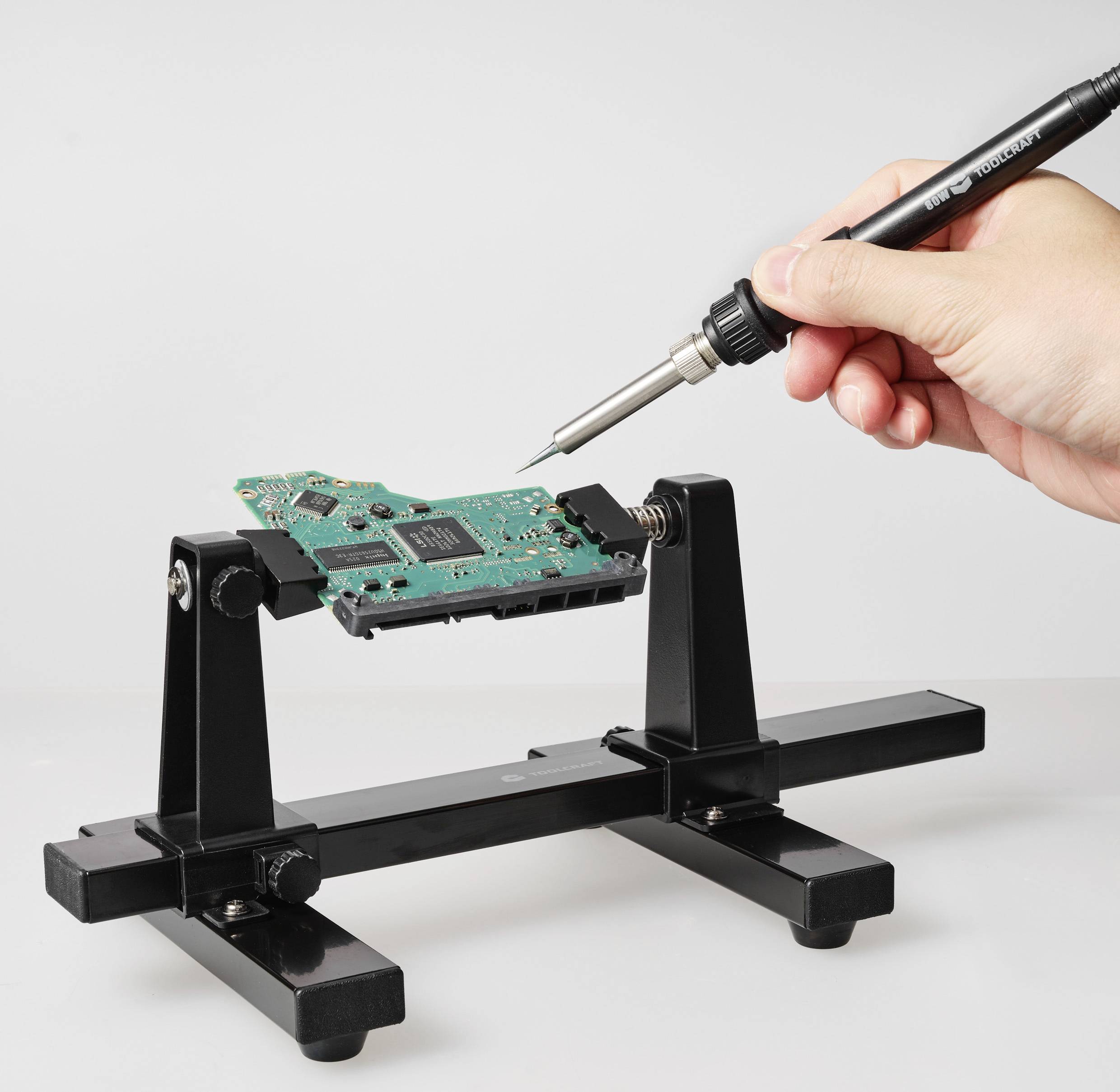 Eine Hand hält einen Lötkolben über eine Leiterplatte, die in einer Halterung fixiert ist.