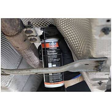 „Liqui Moly Schnell-Rostlöser" kann mit einer behandschuhten Hand auf ein Metallteil unter einem Fahrzeug gesprüht werden, möglicherweise zur Rostentfernung.