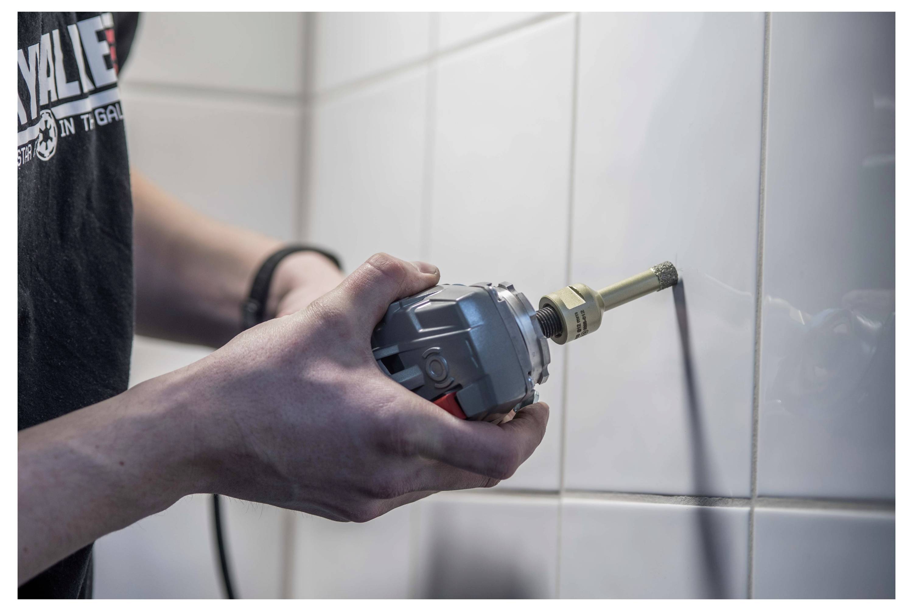 Person, die ein Elektrowerkzeug mit einem Fugen-Reinigungsaufsatz verwendet, um Material aus den Fugen einer weiß gefliesten Badezimmerwand zu reinigen oder zu entfernen.