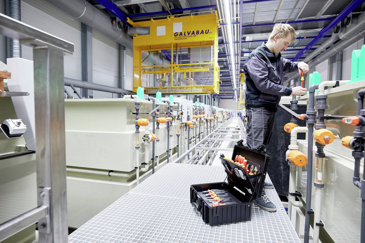 Ein Techniker arbeitet in einer industriellen Anlage mit Maschinen. Vor ihm liegt ein Werkzeugkasten. Die Umgebung ist modern und gut beleuchtet.