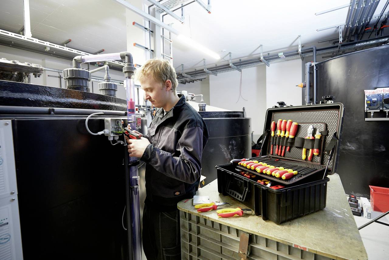 Ein Techniker repariert eine Maschine in einem Wartungsraum. Auf einem Tisch liegt ein offener Werkzeugkoffer mit Schraubendrehern.