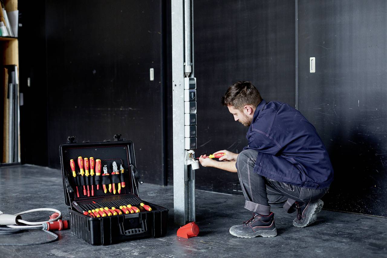Ein Handwerker repariert eine Steckdose in einem Industriegebäude. Neben ihm liegt ein offener Werkzeugkoffer mit diversen Schraubenziehern.