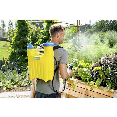 Eine Person mit einer gelben Rückenspritze bewässert Gemüse in einem Garten. Im Hintergrund sind hohe Pflanzen zu sehen.