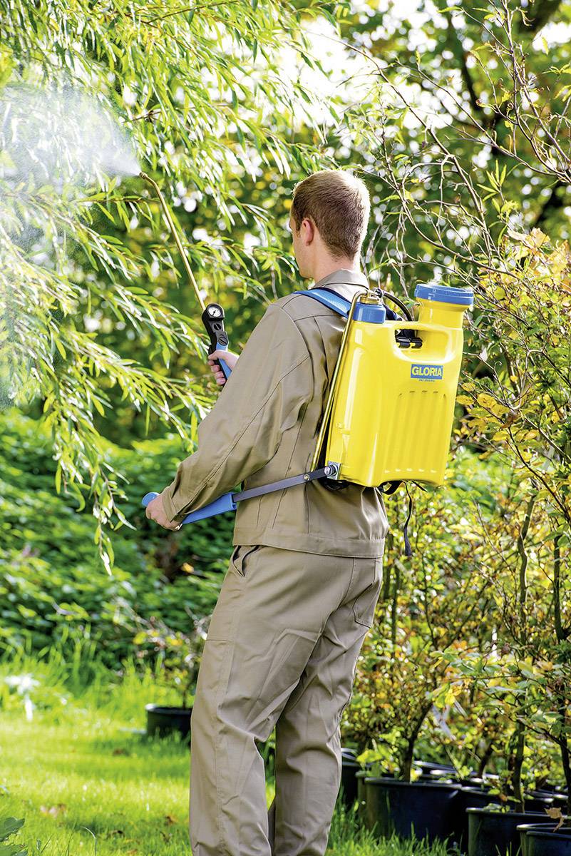 Eine Person in beiger Arbeitskleidung sprüht Pflanzen im Freien mit einem gelben Rückensprühgerät. Sie steht in einem üppigen Garten.