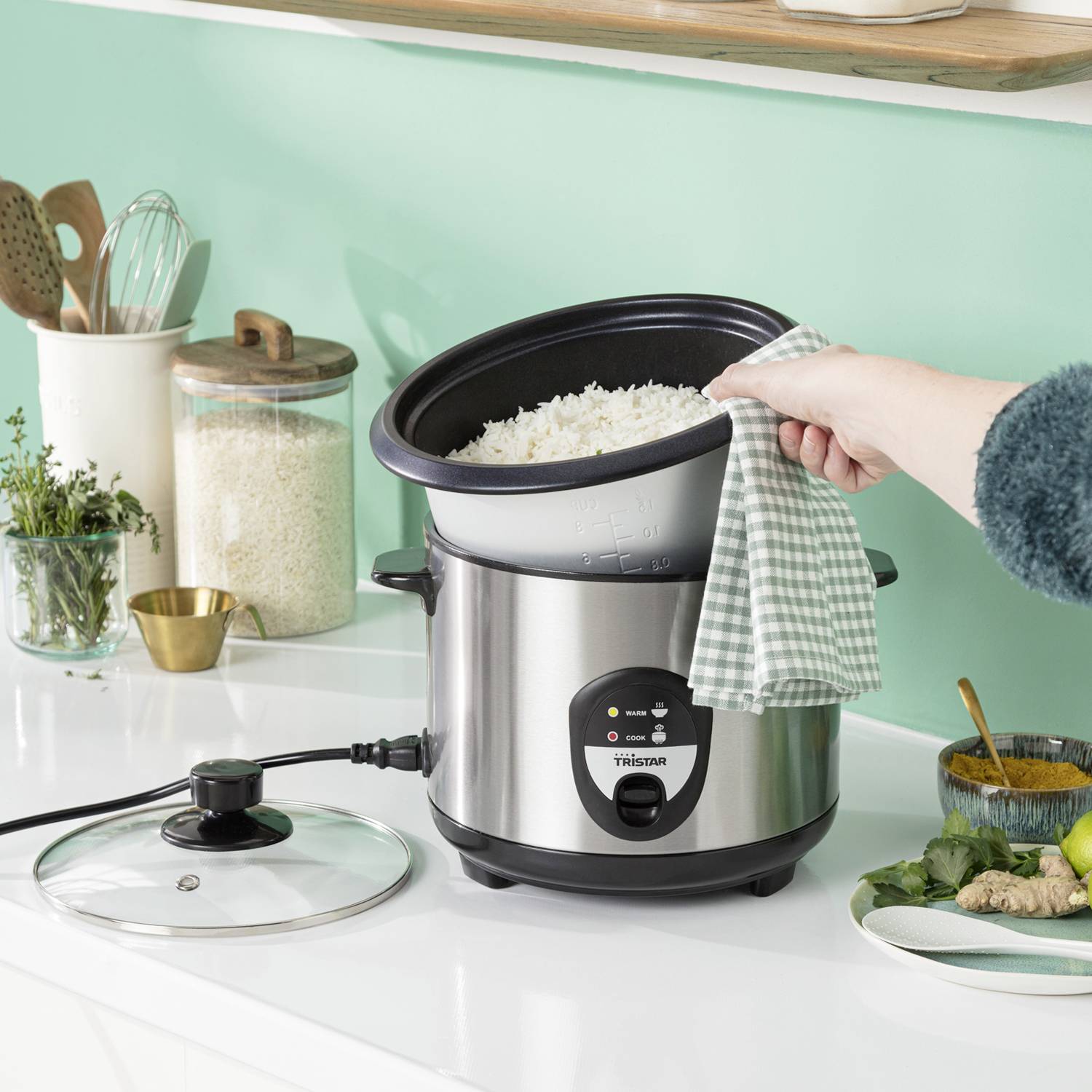 Eine Person hebt den Deckel eines elektrischen Reiskochers, der Reis enthält, in einer Küche mit grüner Wand und Utensilien.