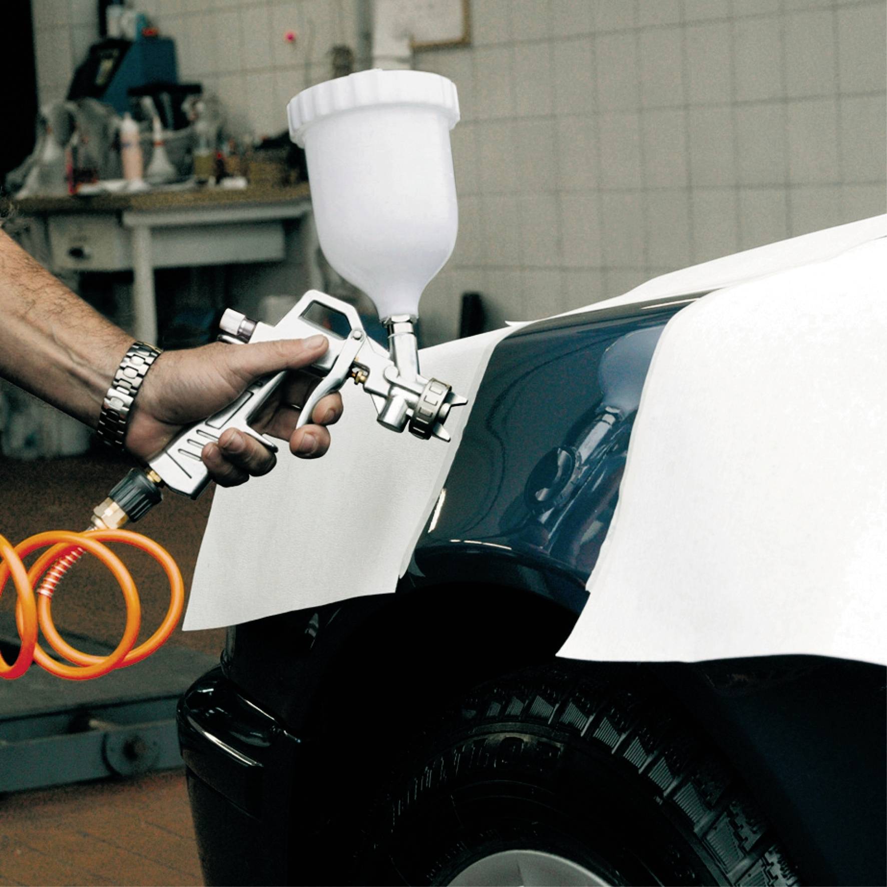 Eine Person lackiert den Kotflügel eines Autos mit einer Spritzpistole. Die Farbe erscheint glänzend. Im Hintergrund ist eine Werkstatt.