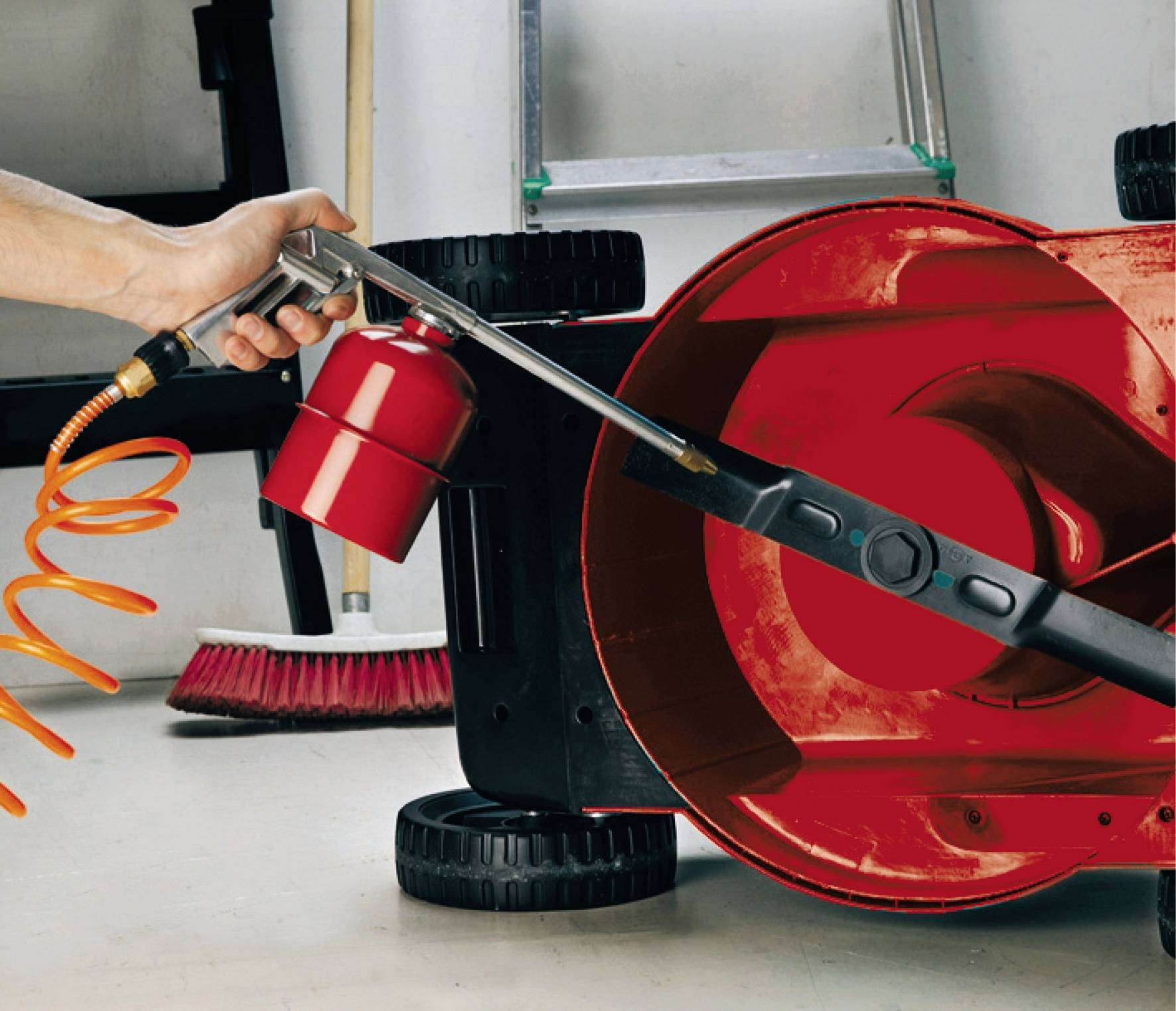 Hand verwendet Druckluftreiniger, um das Gehäuse eines umgedrehten Rasenmähers zu reinigen. Im Hintergrund ist ein Besen zu sehen.