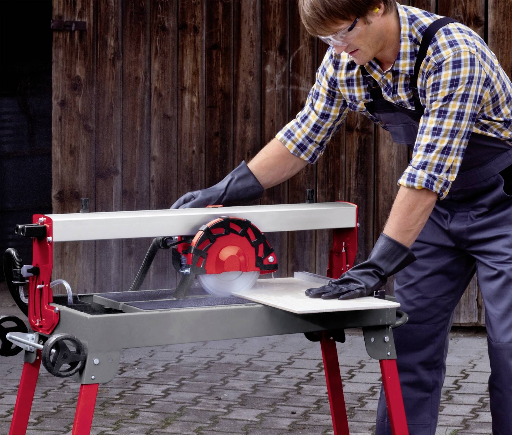 Ein Mann mit Schutzbrille und Handschuhen schneidet ein Brett mit einer Tischkreissäge draußen vor einem Holzwandhintergrund.
