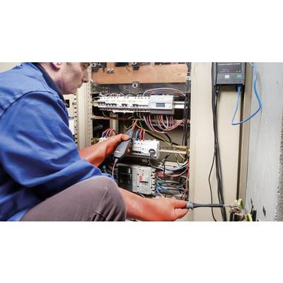 Ein Elektriker in blauer Arbeitskleidung und orange Handschuhen überprüft mit einem Gerät ein offenes Schaltschrank voller Kabel.