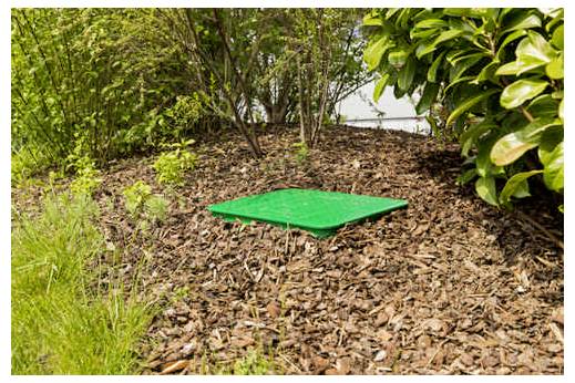 Grüne Bodenbox, installiert in einem Blumenbeet, umgeben von Mulch und Pflanzen.