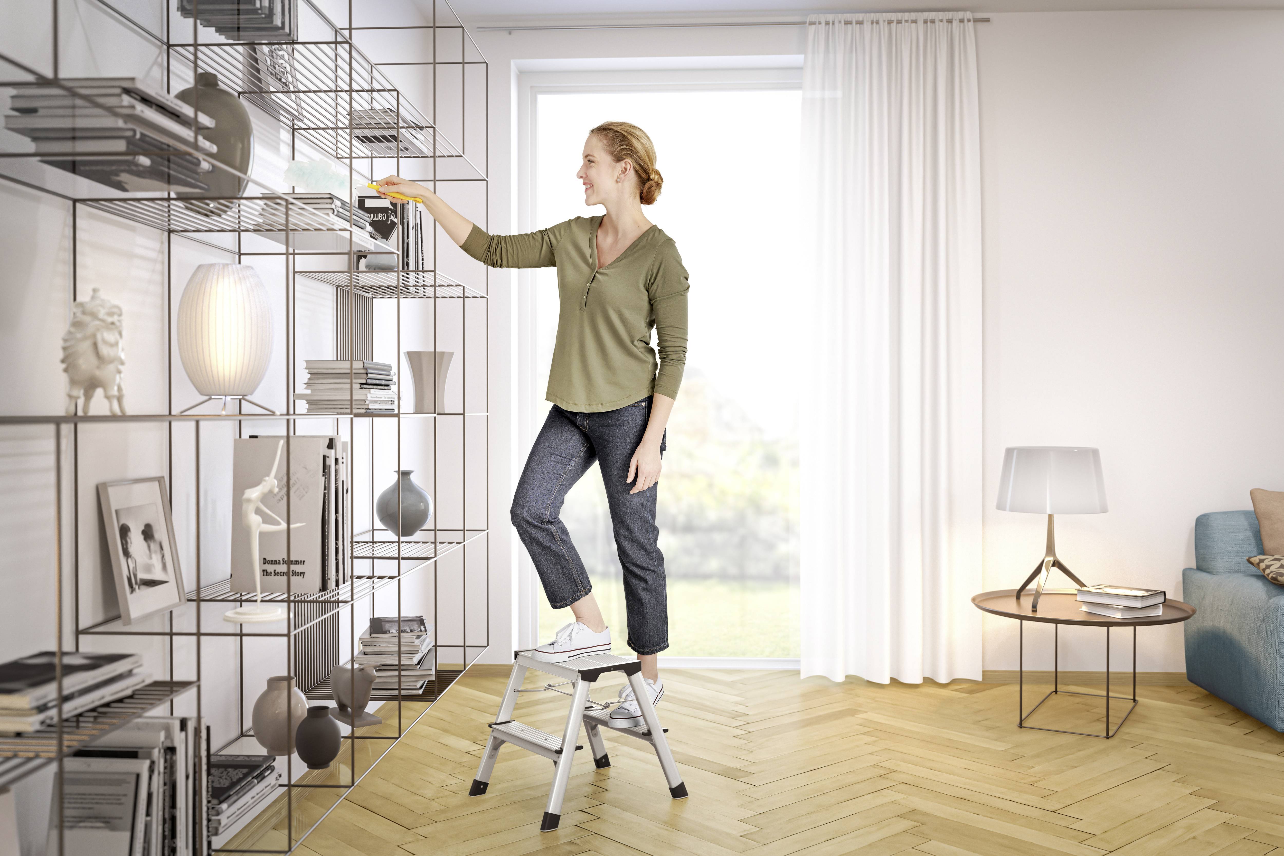 Eine Frau steht auf einer kleinen Leiter und arrangiert Bücher in einem modernen Wohnzimmerregal.