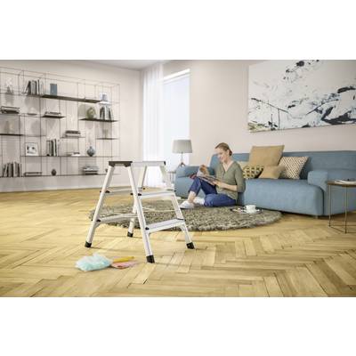 Eine Person sitzt auf einem blauen Sofa in einem Wohnzimmer, liest ein Buch. Im Vordergrund steht eine kleine Leiter.