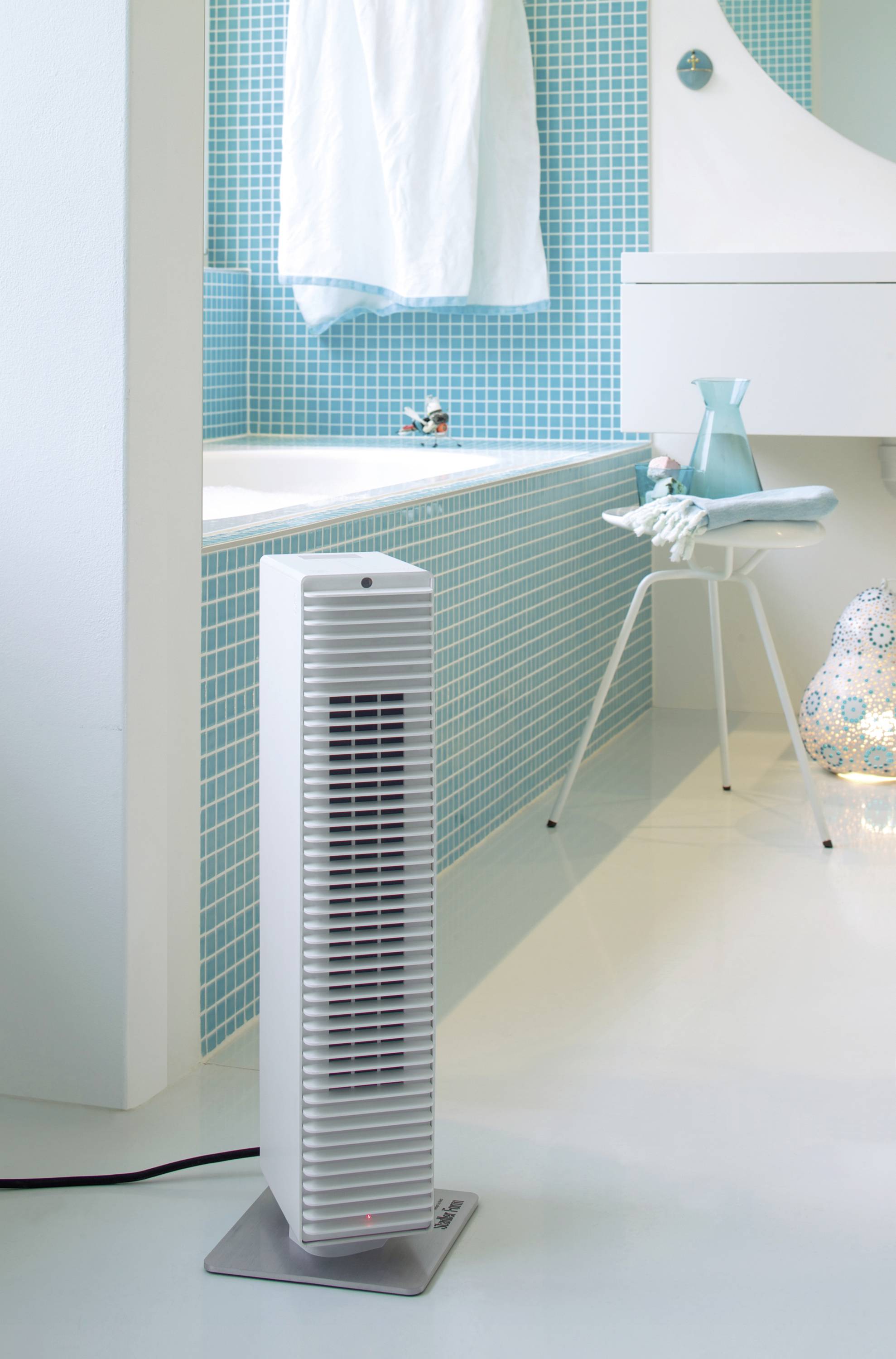 Ein schlanker, weißer Turmventilator steht in einem modernen Badezimmer mit blauen Mosaikfliesen, neben einer weißen Badewanne.