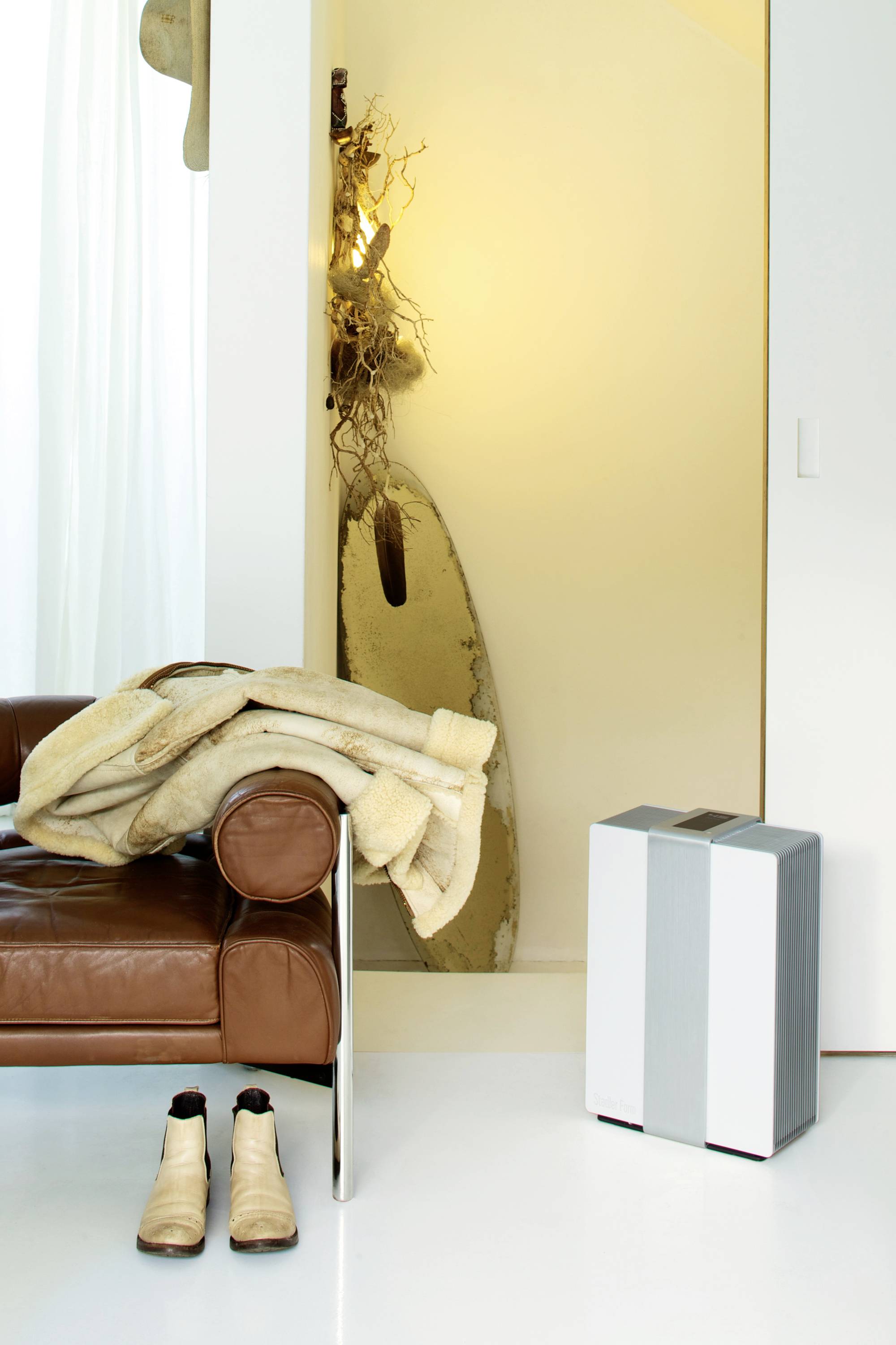 Ein modern eingerichtetes Wohnzimmer mit Ledersessel, Felljacke, abgestellten Stiefeln und einem Luftreiniger auf weißem Boden.