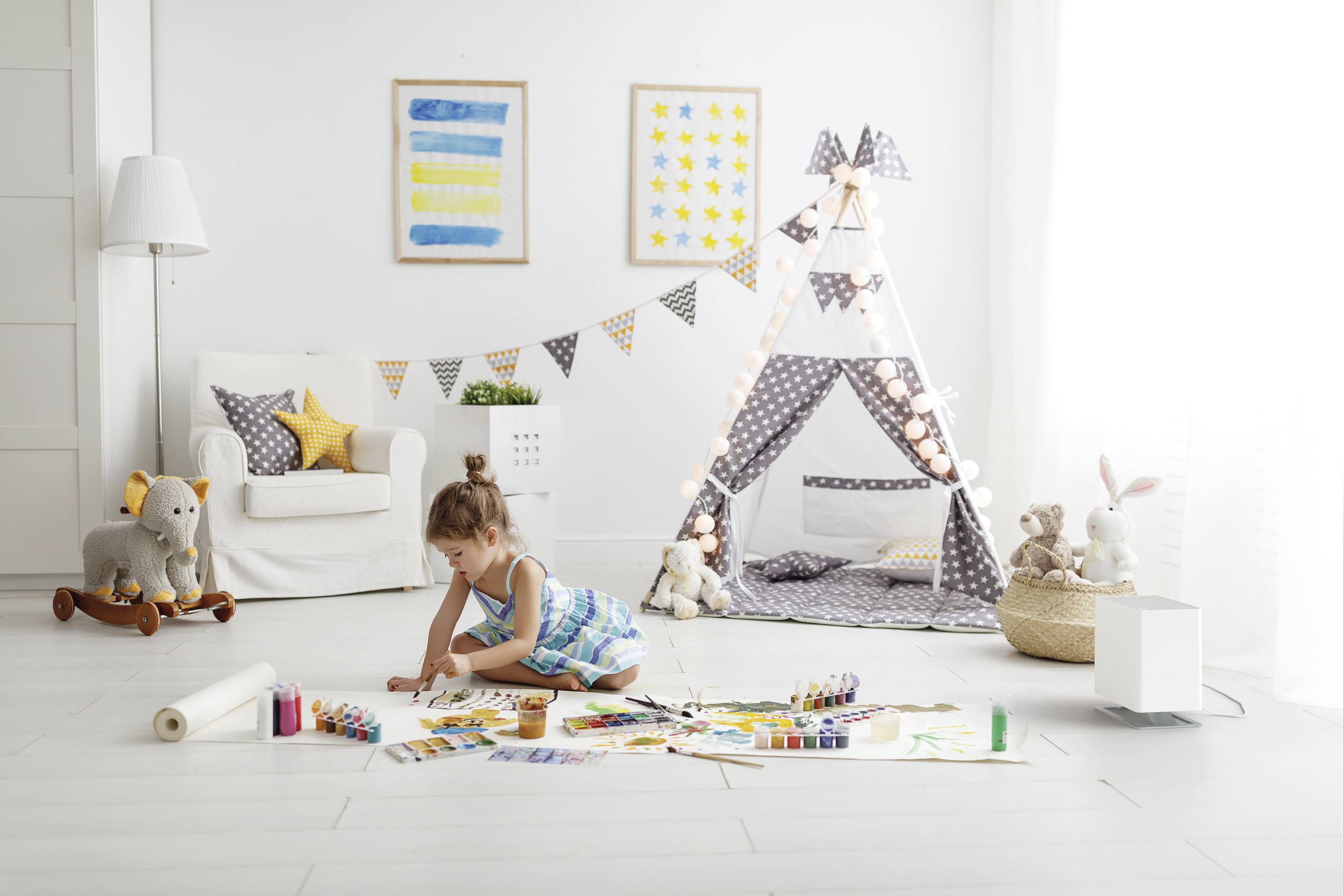 Ein kleines Mädchen malt auf dem Boden eines hellen Kinderzimmers, umgeben von Spielzeug und einem dekorativen Tipi-Zelt.