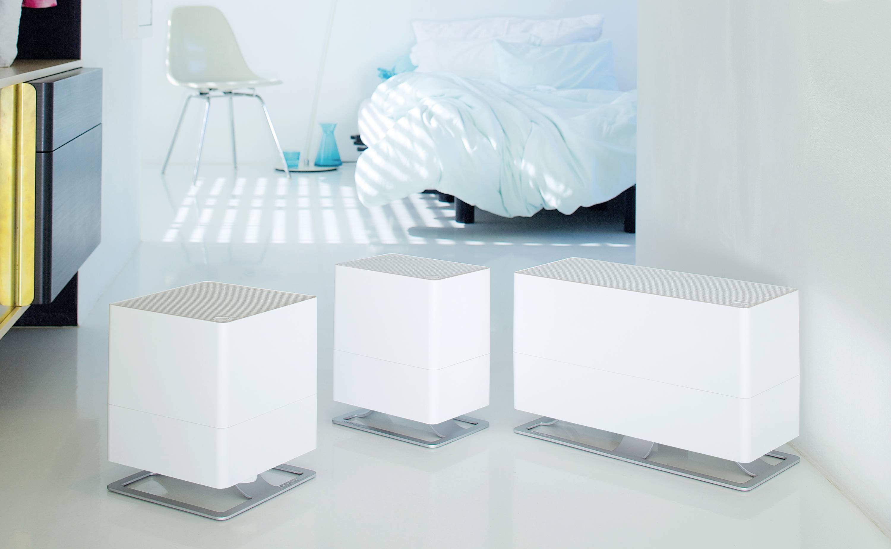 Drei moderne weiße Luftbefeuchter unterschiedlicher Größe stehen in einem stilvollen, minimalistisch eingerichteten Raum mit Bett im Hintergrund.