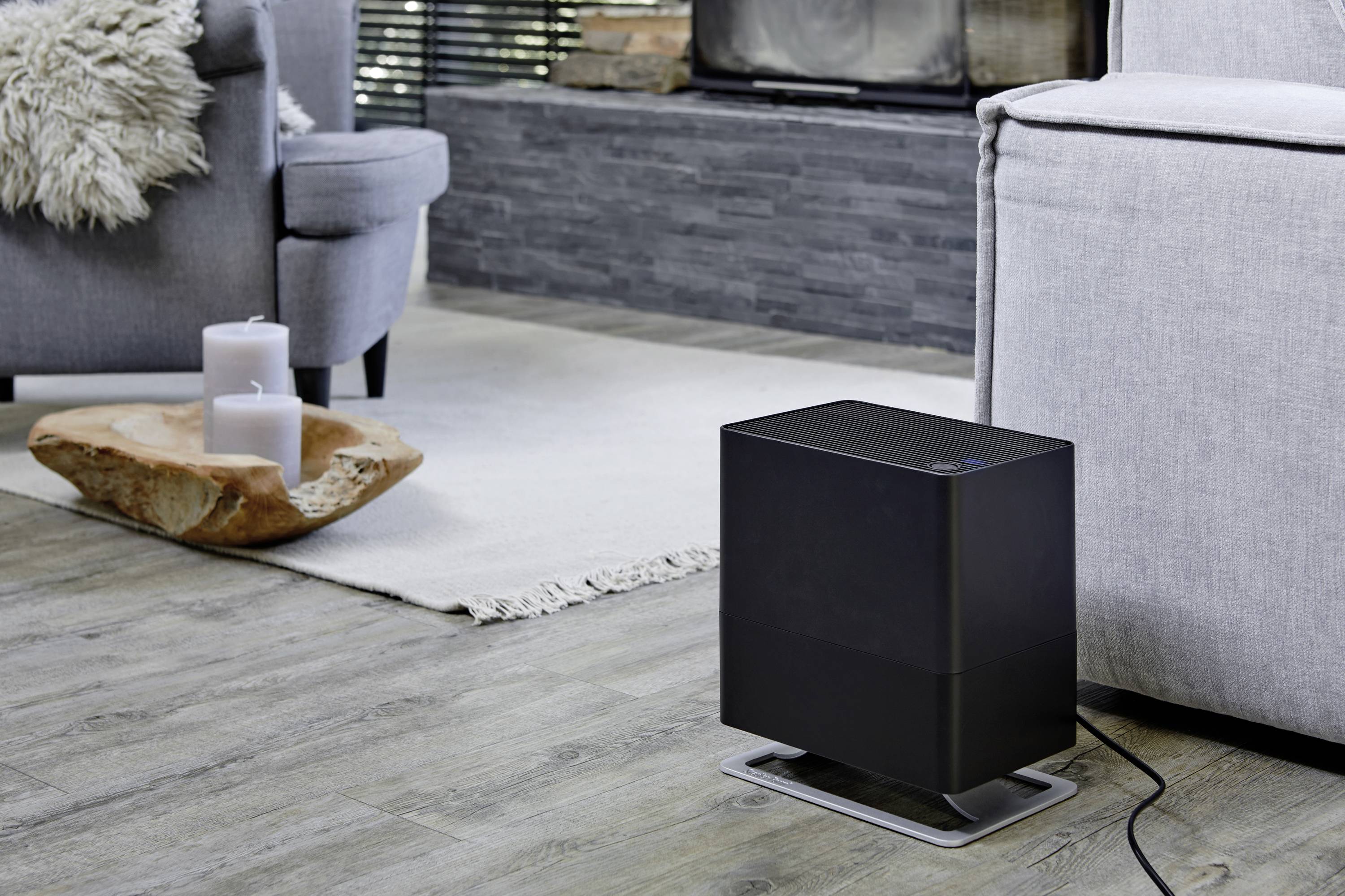 Ein schwarzer Heizlüfter steht auf einem Holzboden neben einem grauen Sofa in einem modernen Wohnzimmer. Kerzen auf Holzschale daneben.