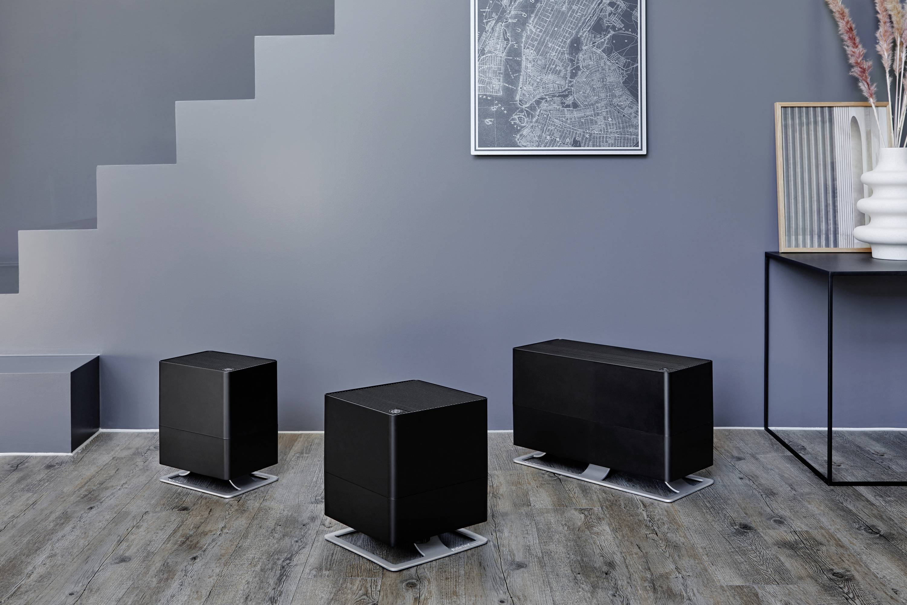 Drei schwarze Luftbefeuchter in verschiedenen Größen auf einem Holzboden in einem modernen Raum mit grauer Wand und minimaler Dekoration.