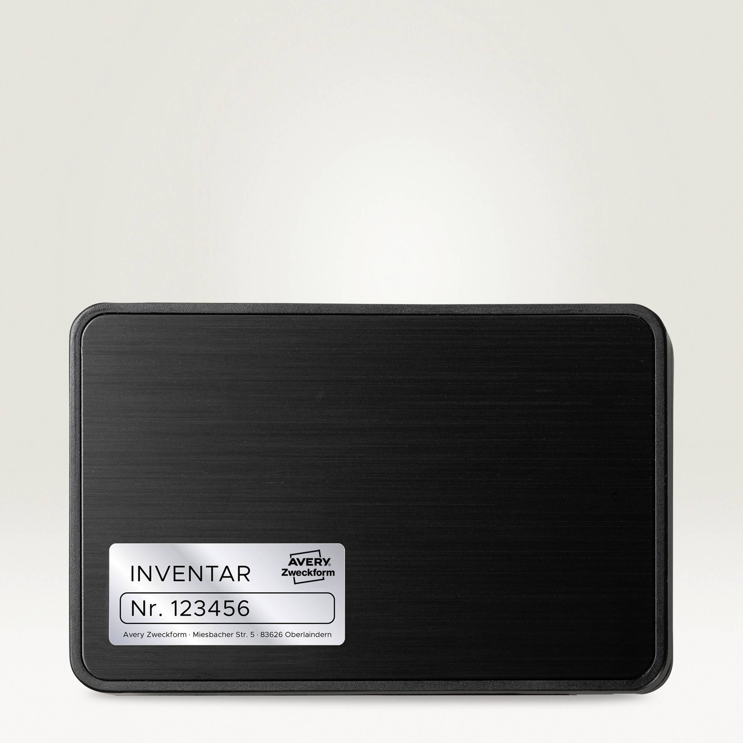 Schwarzes Schild mit weißem Etikett: 'INVENTAR Nr. 123456', darunter 'Avery Zweckform'.