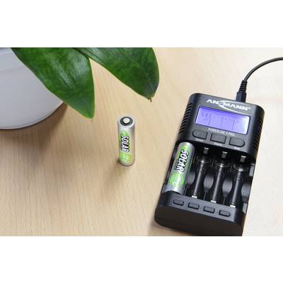 Ein Batterieladegerät mit vier leeren Ladeschächten liegt auf einem Tisch neben einer Pflanze. Eine einzelne AA-Batterie steht daneben.