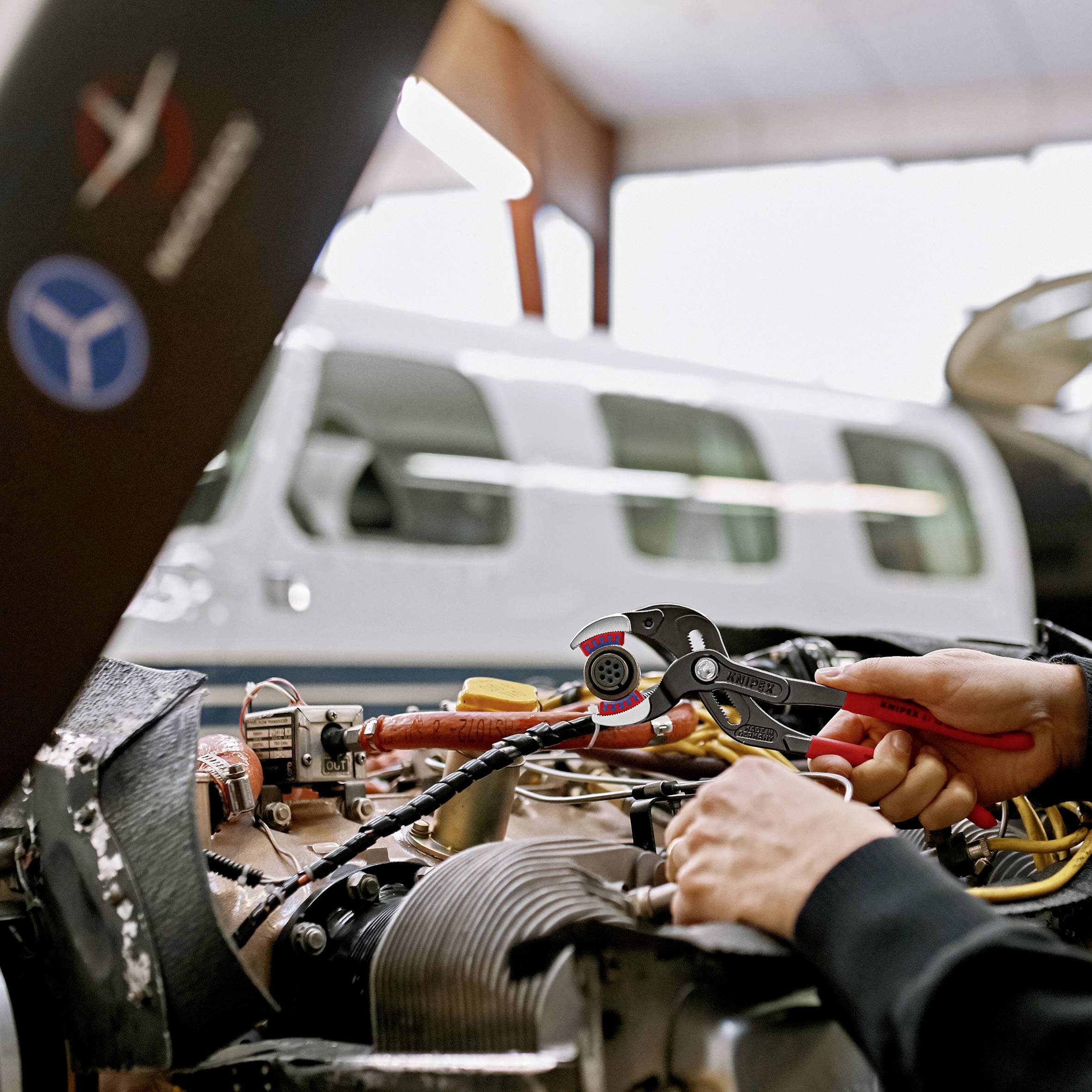 Ein Techniker repariert einen Flugzeugmotor mit Werkzeugen. Im Hintergrund steht ein Flugzeug im Hangar.