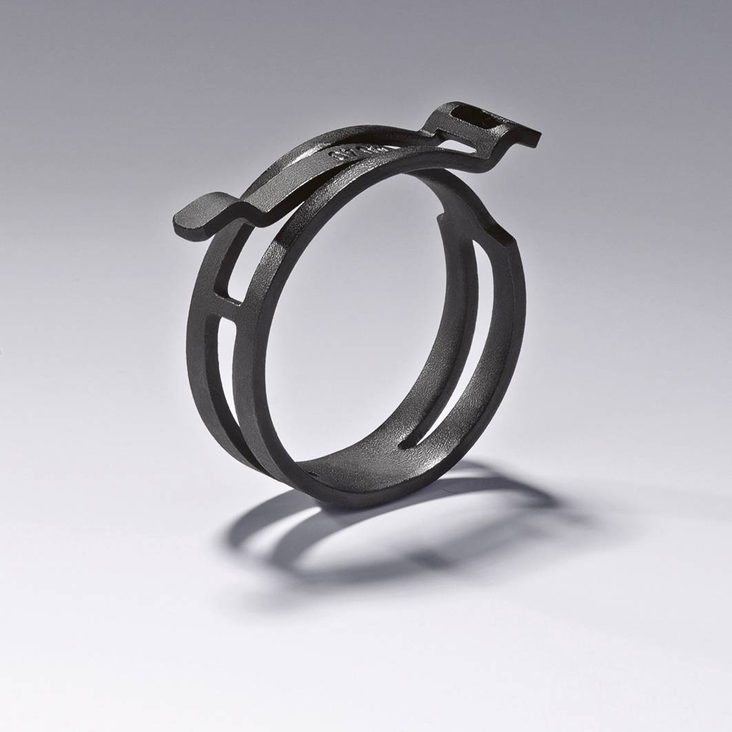 Ein schwarzer Metallring mit asymmetrischem Design auf einer grauen Oberfläche. Der Ring wirft einen Schatten nach rechts.