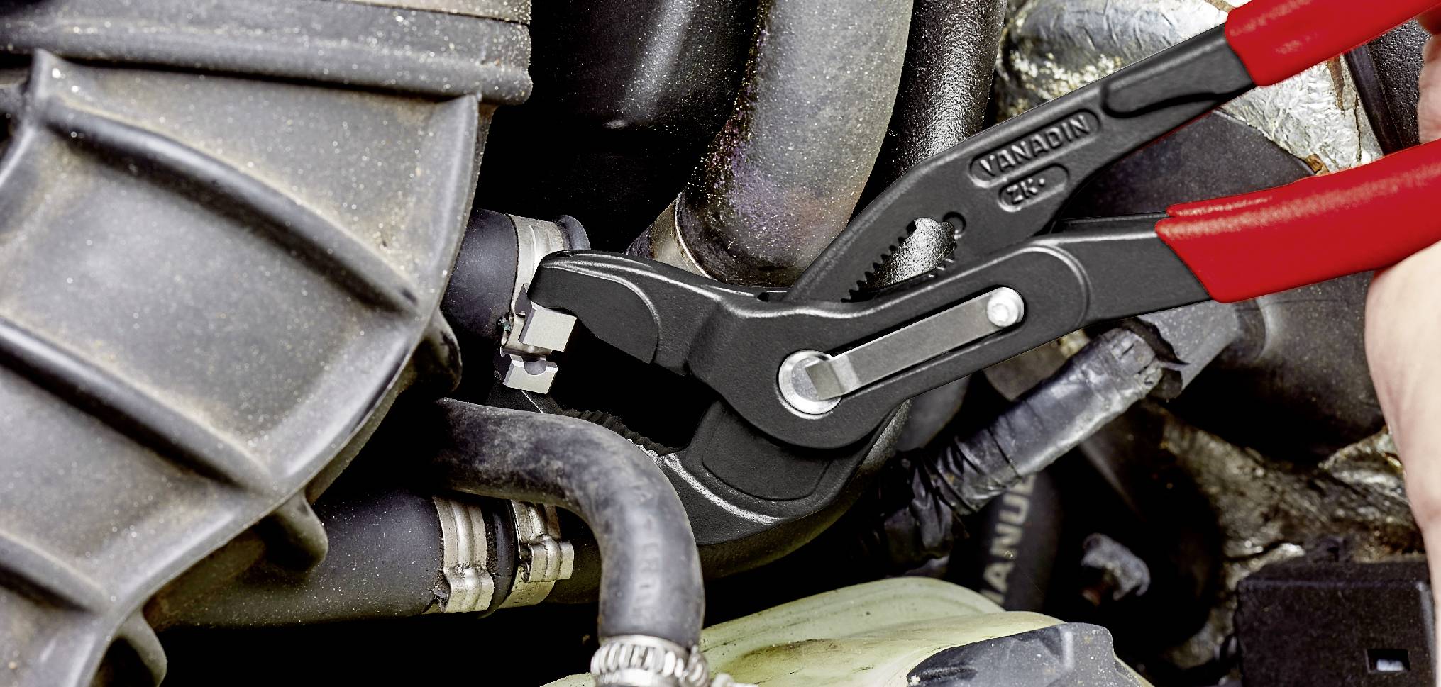 Eine Hand hält eine Zange, die an einem Autoteil arbeitet. Die Zange greift eine Schlauchverbindung im Motorraum.