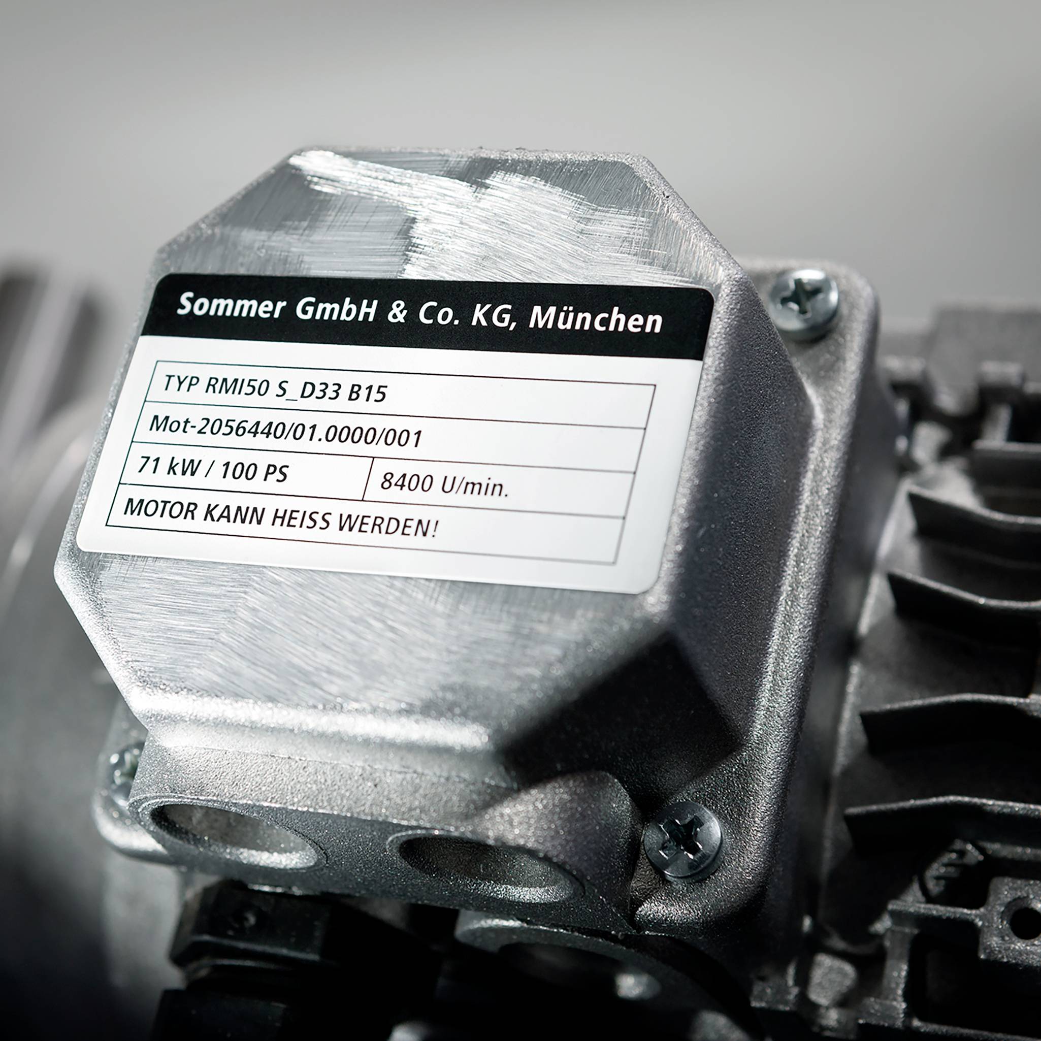 Etikett auf einem Metallgehäuse mit Informationen: 'Sommer GmbH & Co. KG, München. Typ RMI50 S_D33 B15, 71 kW, 400 V/1000 P008, 8400 U/min. MOTOR KANN HEISS WERDEN!'.