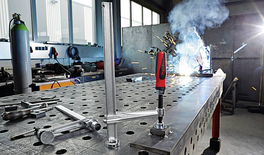 Ein Tisch mit Metallwerkzeugen und einer Vorrichtung zum Schweißen; im Hintergrund schweißt eine Person funkenreich Metall.