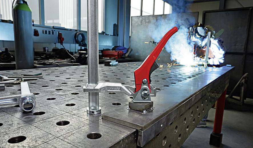 Ein Schweißer arbeitet an einem Metalltisch mit einem roten Schraubzwinge im Vordergrund. Funken fliegen während des Schweißvorgangs.