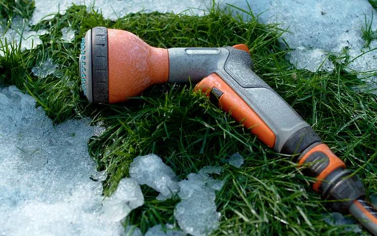 Eine orange-graue Gartenschlauchbrause liegt auf grünem Gras, umgeben von Stellen mit schmelzendem Eis und Schnee, was den frühen Frühling andeutet.