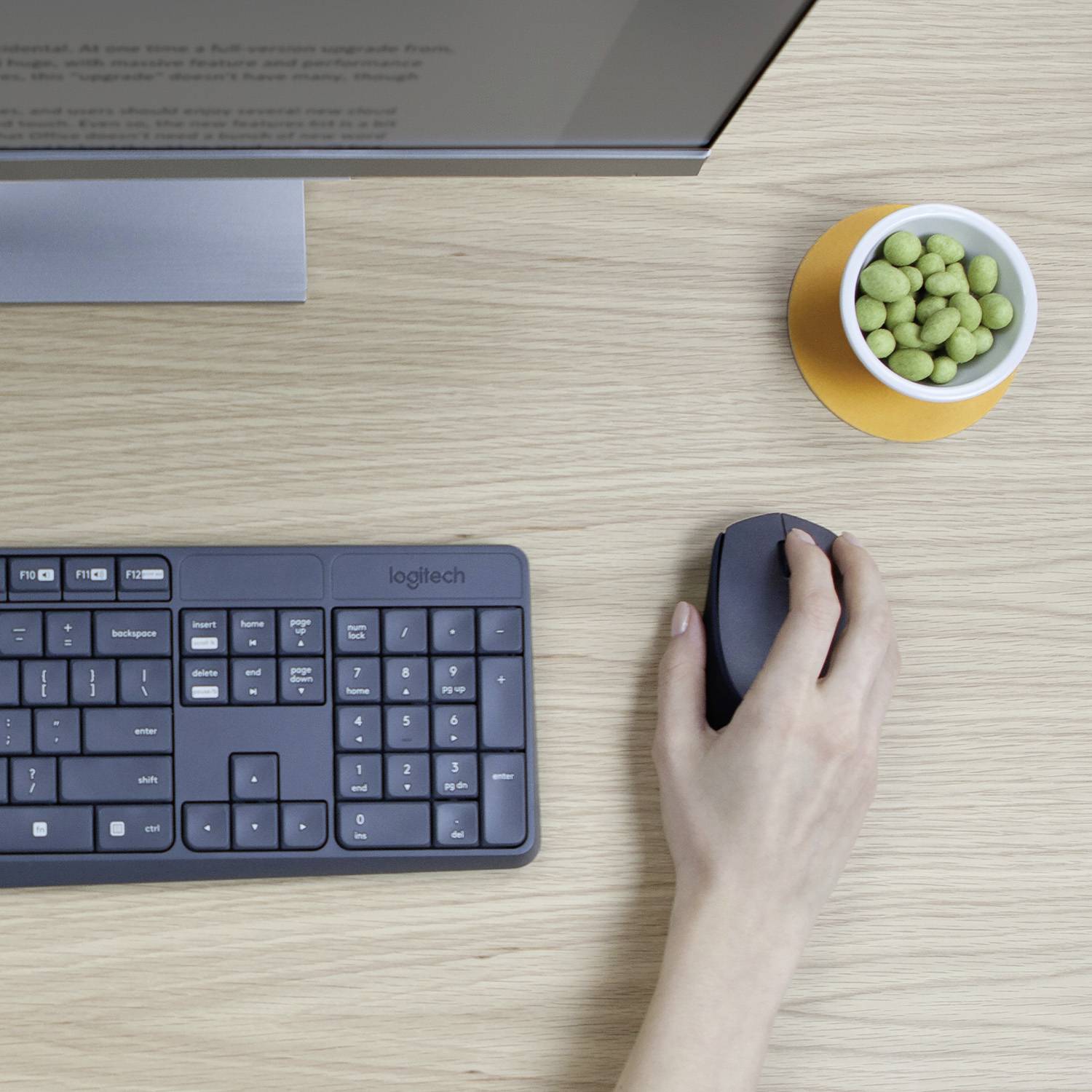 Eine Person benutzt eine kabellose Maus neben einer Tastatur auf einem Schreibtisch. Daneben steht eine Schale mit grünen Snacks.