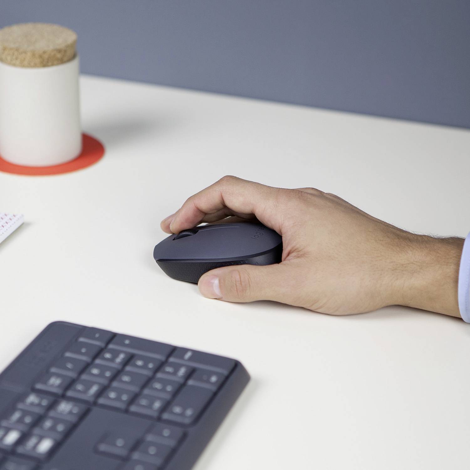 Eine Hand hält eine Computermaus auf einem weißen Tisch neben einer Tastatur. Im Hintergrund steht ein Becher auf einem roten Untersetzer.