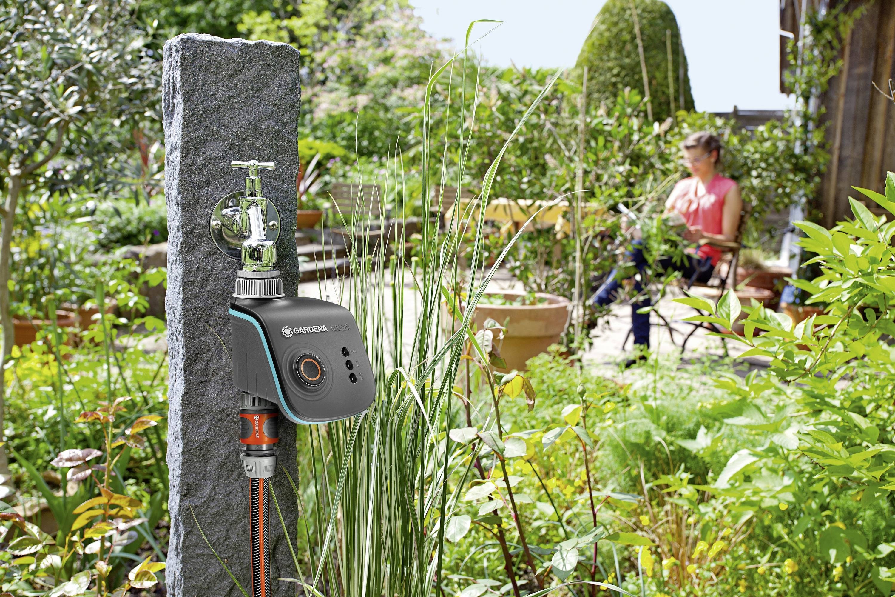 Eine automatische Gartenschlauchsteuerung an einem Wasserhahn im Vordergrund. Im Hintergrund sitzt eine Person in einem grünen Garten.