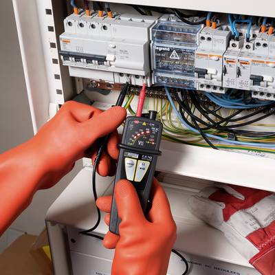 Elektriker in orangefarbenen Handschuhen prüft mit einem Multimeter die Spannungsversorgung in einem geöffneten Schaltschrank.