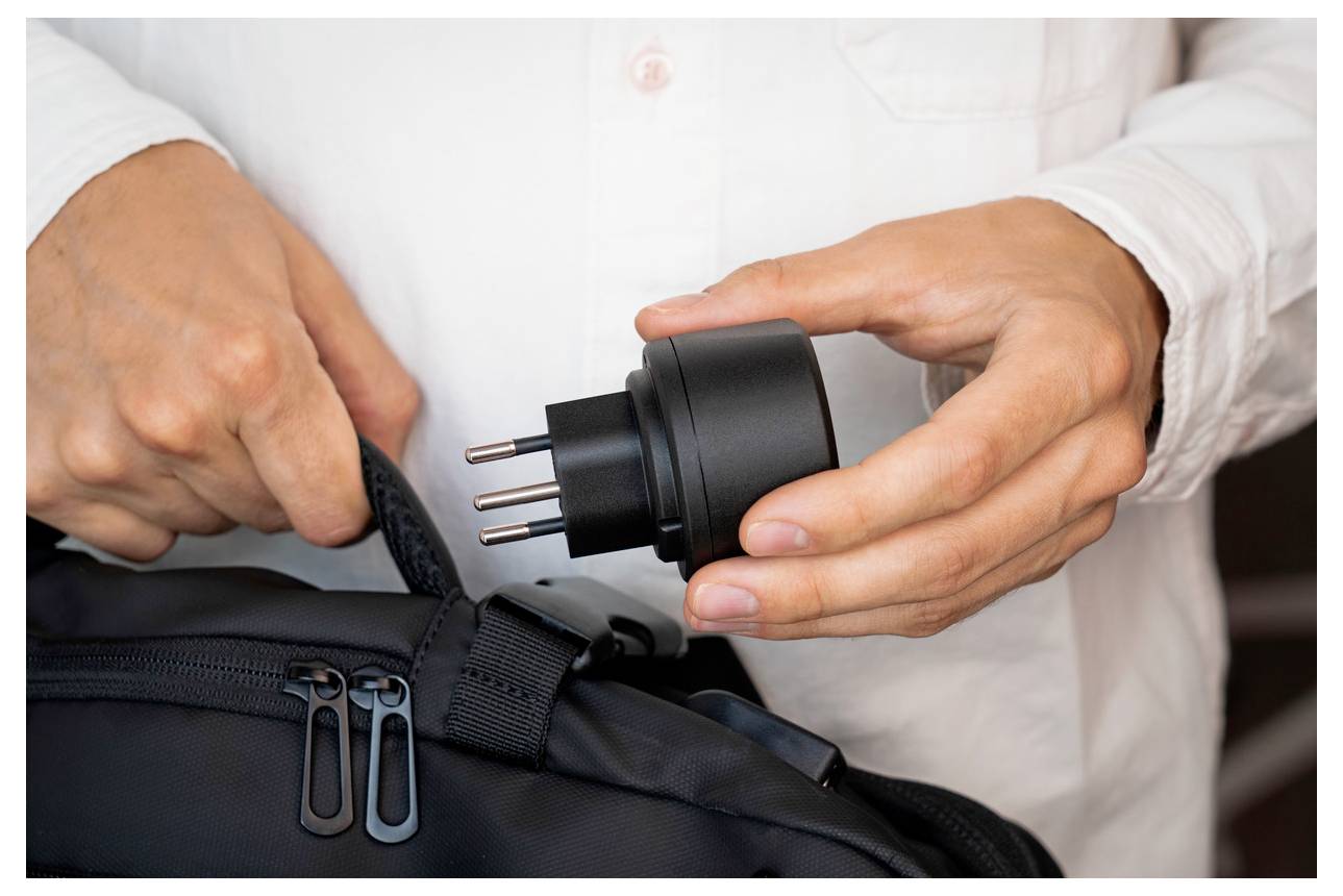 Person packt eine schwarze Reisetasche mit einem dreipoligen Stromstecker oder Adapter.