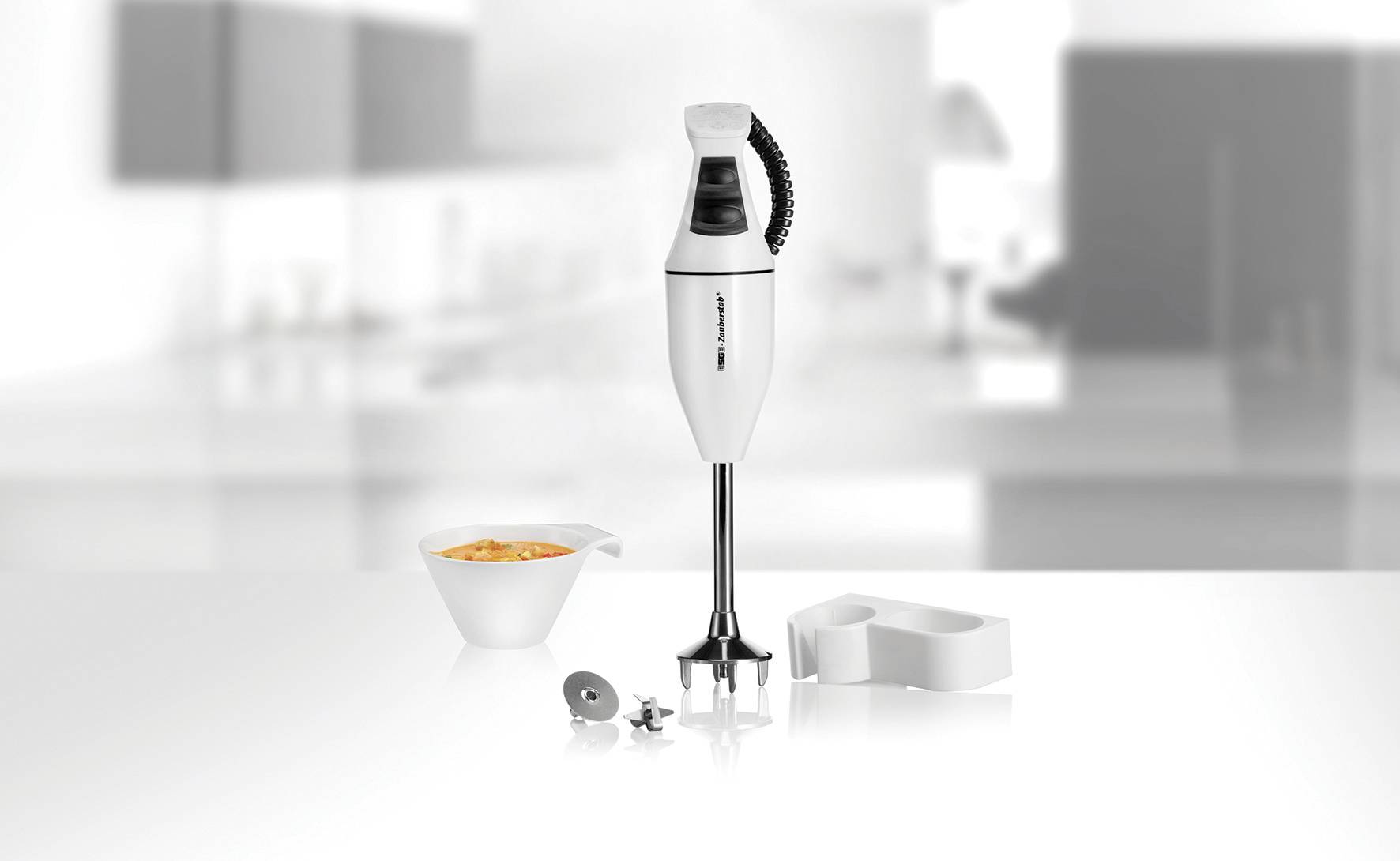 Ein weißer Stabmixer steht auf einer Küchenarbeitsplatte, daneben eine Schüssel mit orangefarbenem Inhalt und einige Zubehörteile.