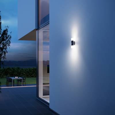 Moderne Architektur bei Dämmerung, beleuchtet von einer Wandleuchte. Tisch und Stühle im Garten sichtbar.