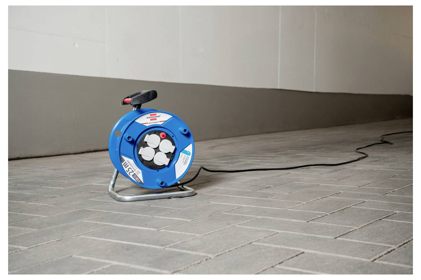 Eine blaue tragbare Kabelrolle mit schwarzem Griff steht auf einem gepflasterten Boden gegen eine neutrale Wand gelehnt.